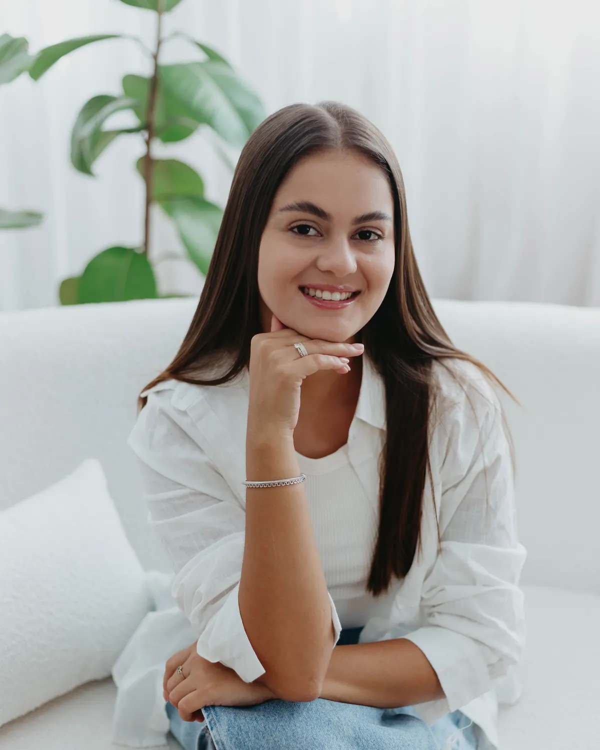 Mikaella Nunes de Souza sentada em um sofá claro, em ambiente com plantas e luz natural